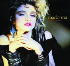 Front cover of Madonna’s 1983 Italian LP “The First Album”, featuring her iconic early-80s look with blonde curls, cross earring, layered jewelry, and bold makeup, photographed by Gary Heery for the Sire Records release.